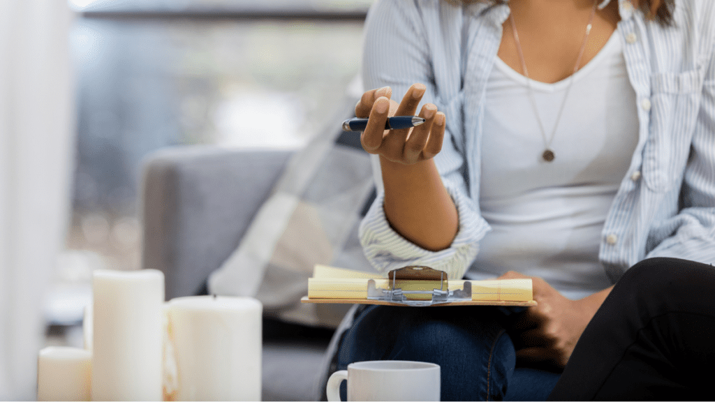 A therapist or mental health practitioner seated in a therapy room, holding a pen and clipboard with notes, in a calm and informal clinical setting.